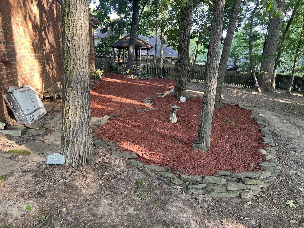 Red mulch bed ringed with stacked stone around tree trunks in a shaded backyard.