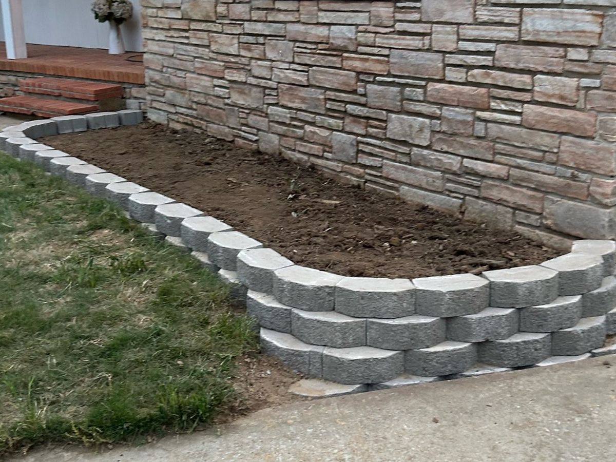 New bed of bare soil framed in stacked retaining block against a stone-faced wall.